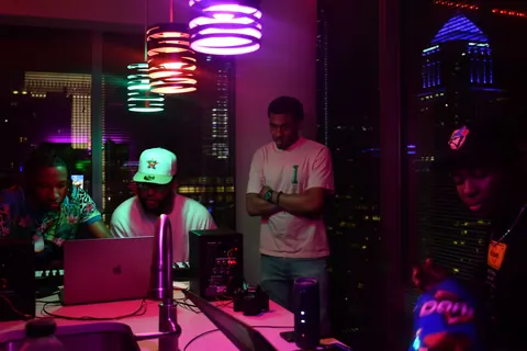A DJ works behind a booth in a club lit with purple and green light.