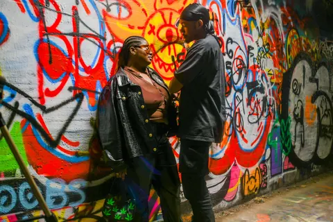 A couple in dark clothing stands close together before a graffiti-covered wall.