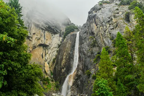 A tall waterfall drops through a granite valley framed by green trees.