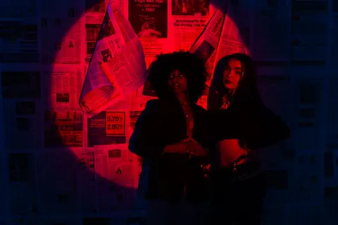 Two people stand in red and blue light against a wall covered in newspaper pages.