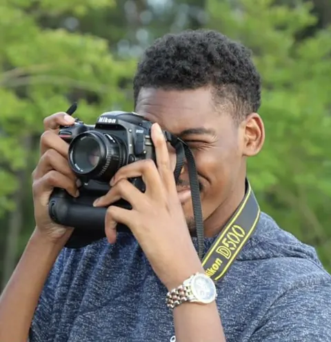 Joshua McKenzie German raising a camera to his eye outdoors.