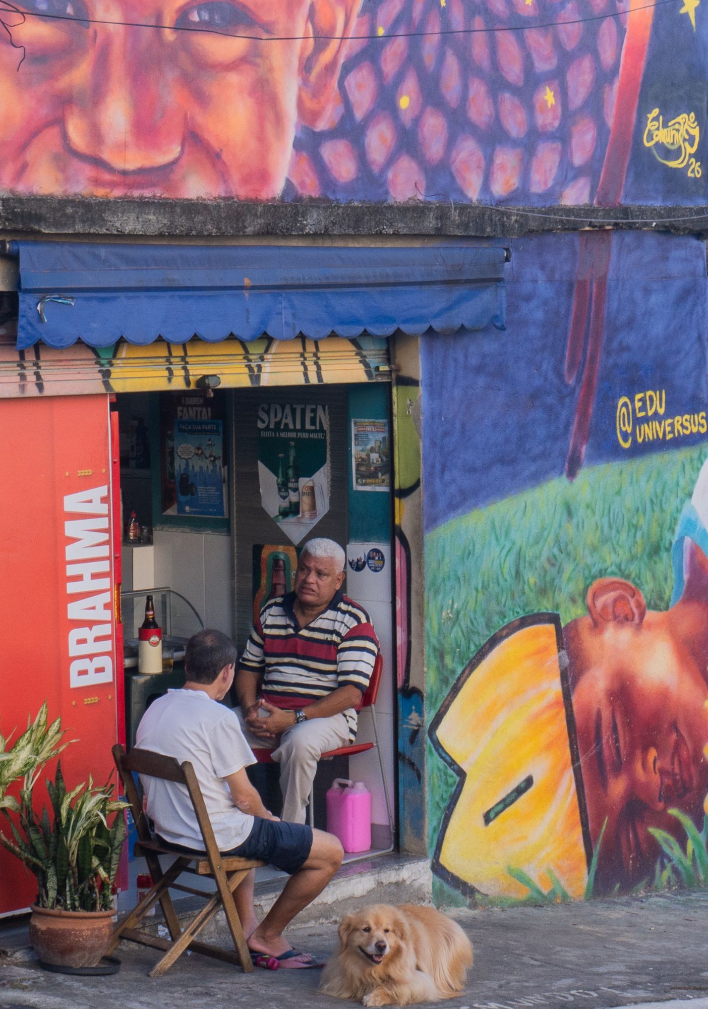 Two men share a conversation outside a corner bar in Rio de Janeiro, a golden dog resting at their feet, a mural of a painted face on the wall behind them.